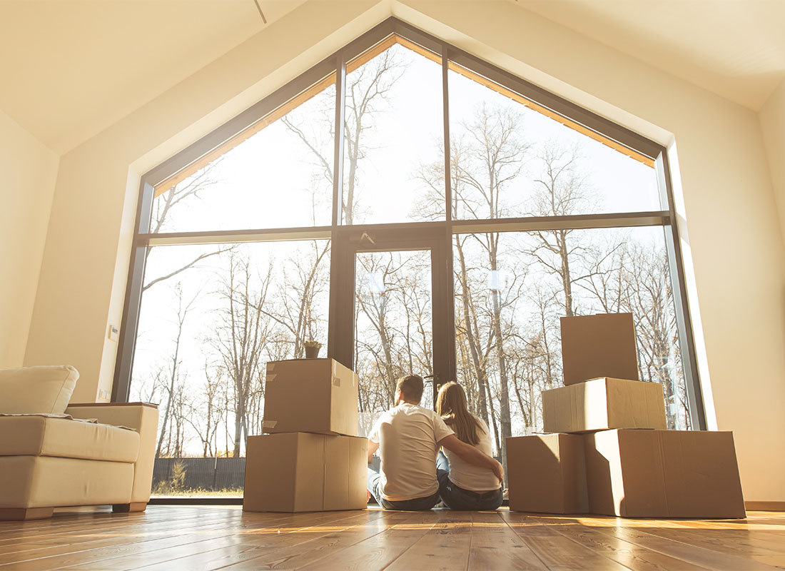 Insurance Solutions - Rear View of a Young Married Couple Sitting on the Floor in Their New Home Next to Moving Boxes Looking Out the Full Length Windows