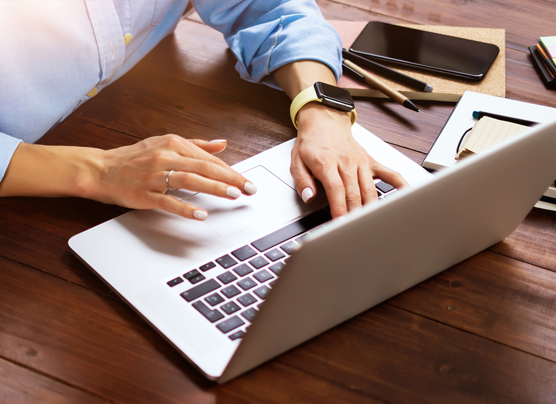 Educational Resources - Woman Typing on an Open Laptop While Sitting at a Wooden Table with Notepads, Her Phone, and Pens on the Table Beside Her